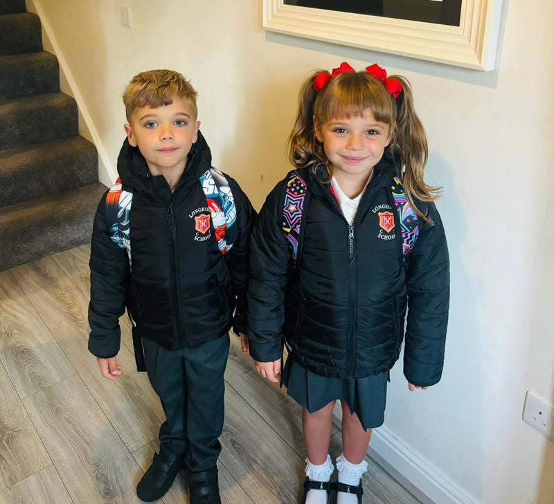Two young children standing indoors wearing matching black puffer jackets with Longridge C of E Primary School crest on the chest printed by Ribble Apparel. The boy wears dark trousers and black shoes and the girl wears a grey skirt, white socks and black shoes. Both have colourful patterned backpacks.