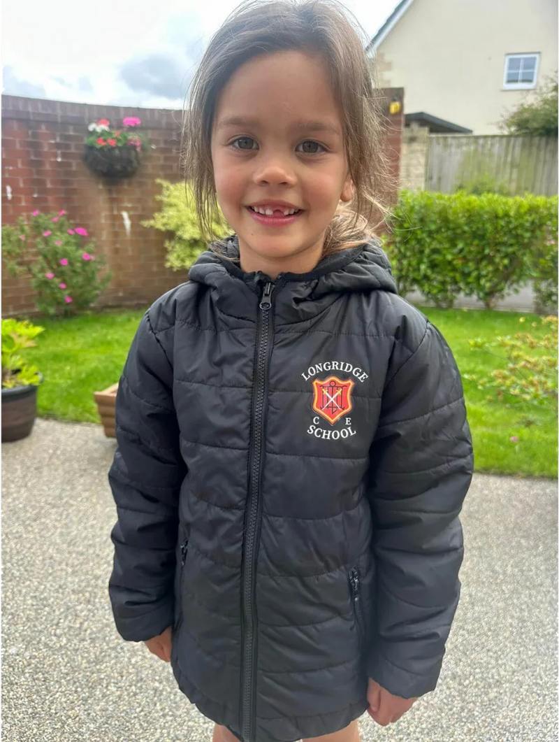 A young girl smiling while wearing a black puffer jacket with Longridge C of E Primary School crest printed on the chest by Ribble Apparel. The photo is taken outdoors in a backyard setting with grass, a brick wall, and a house visible in the background.