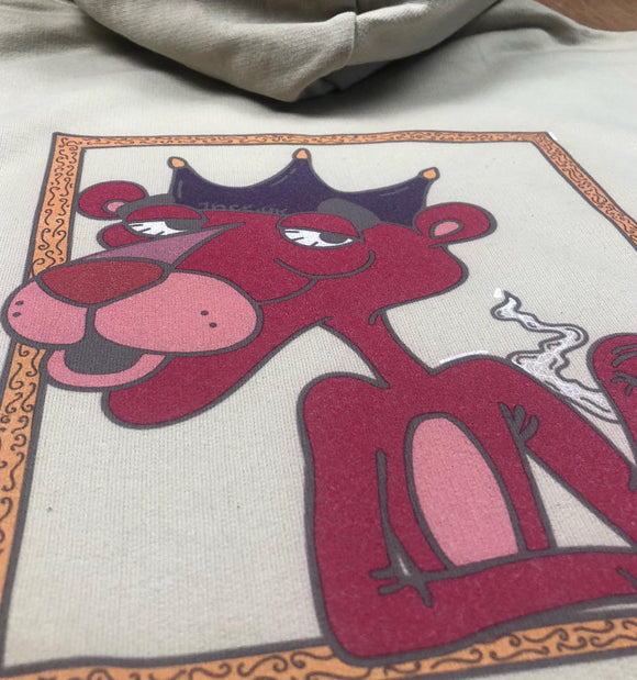 A close-up of a light beige hoodie featuring a graphic print of a red cartoon panther wearing a purple crown and smoking a cigarette.