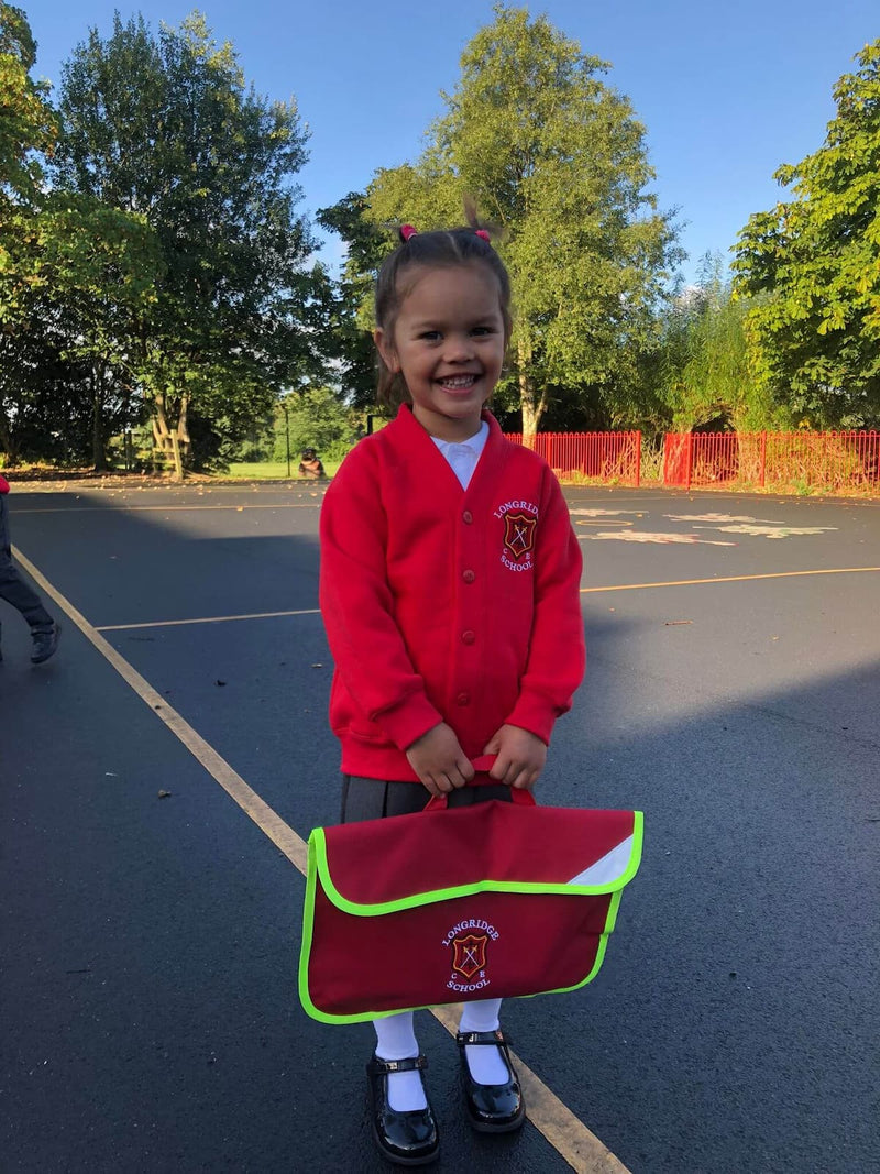 A young girl smiling on an outdoor school playground, wearing Longridge C of E Primary School uniform by Ribble Apparel. She's also holding a matching red and green school bag.