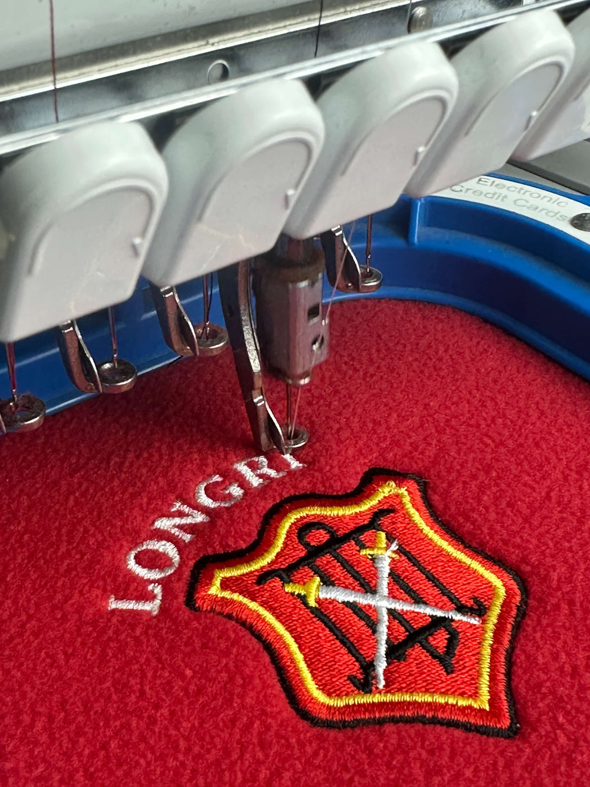 Close-up of an embroidery machine stitching a red embroidered patch with a white cross and black and yellow border, next to white embroidered text on red fabric.