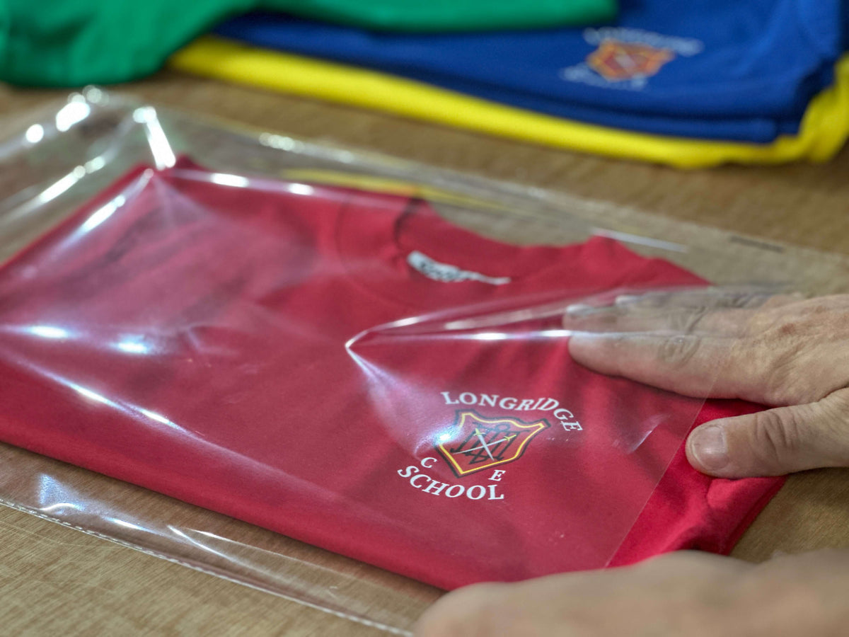 A red folded t-shirt inside a clear plastic sleeve, with a hand smoothing the plastic. The shirt features a 'Longridge C.