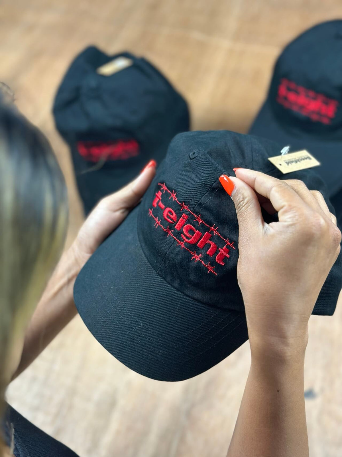 A person holds a black baseball cap with red embroidered text reading 'freight' on the front. Two more matching black caps are blurred in the background on a wood floor.