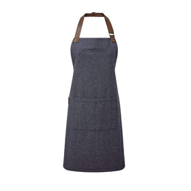 A navy blue bib-apron with a leather-like adjustable strap at the neck and a pen pocket on the front. It is displayed against a white  background. Premier Annex Oxford Bib Apron - PR144