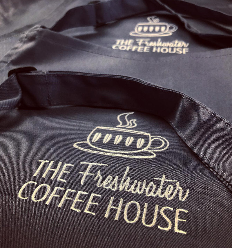 A close-up of a black fabric apron with gold embroidered text reading 'THE Freshwater COFFEE HOUSE' and a coffee cup logo.