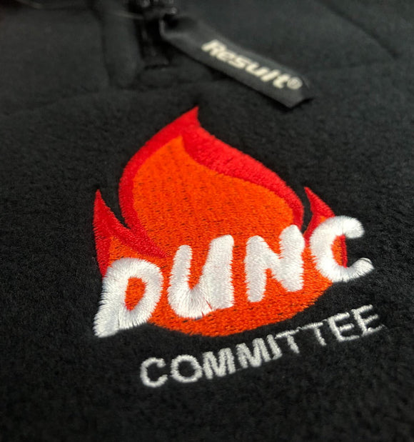 Close-up of a black fleece garment with an embroidered flame logo in red and orange above the white embroidered text 'DUNC COMMITTEE'. A small black hangtag with white text is visible near the zipper.