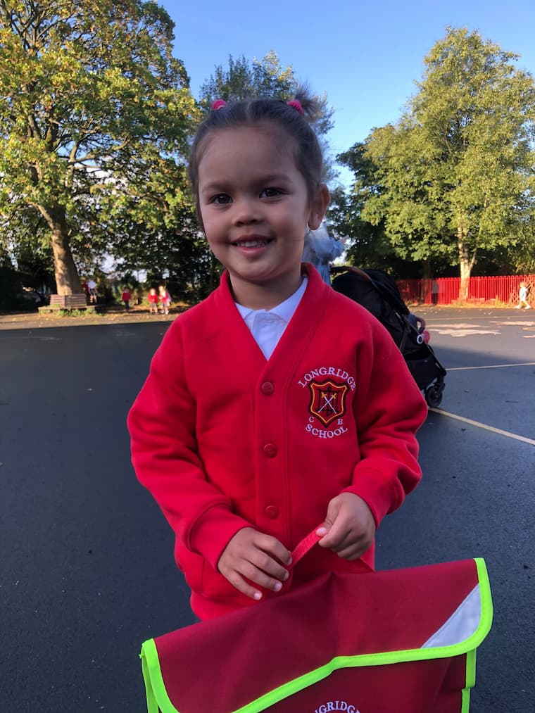 A young child wearing a red button-up cardigan with Longridge C of E School crest by Ribble Apparel standing outdoors in a playground holding a red bag with a bright yellow trim.