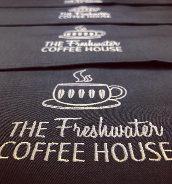 Close-up of a black fabric item, likely a coffee cup coaster or napkin, embroidered with a white coffee cup graphic and the text 'THE Freshwater COFFEE HOUSE'.