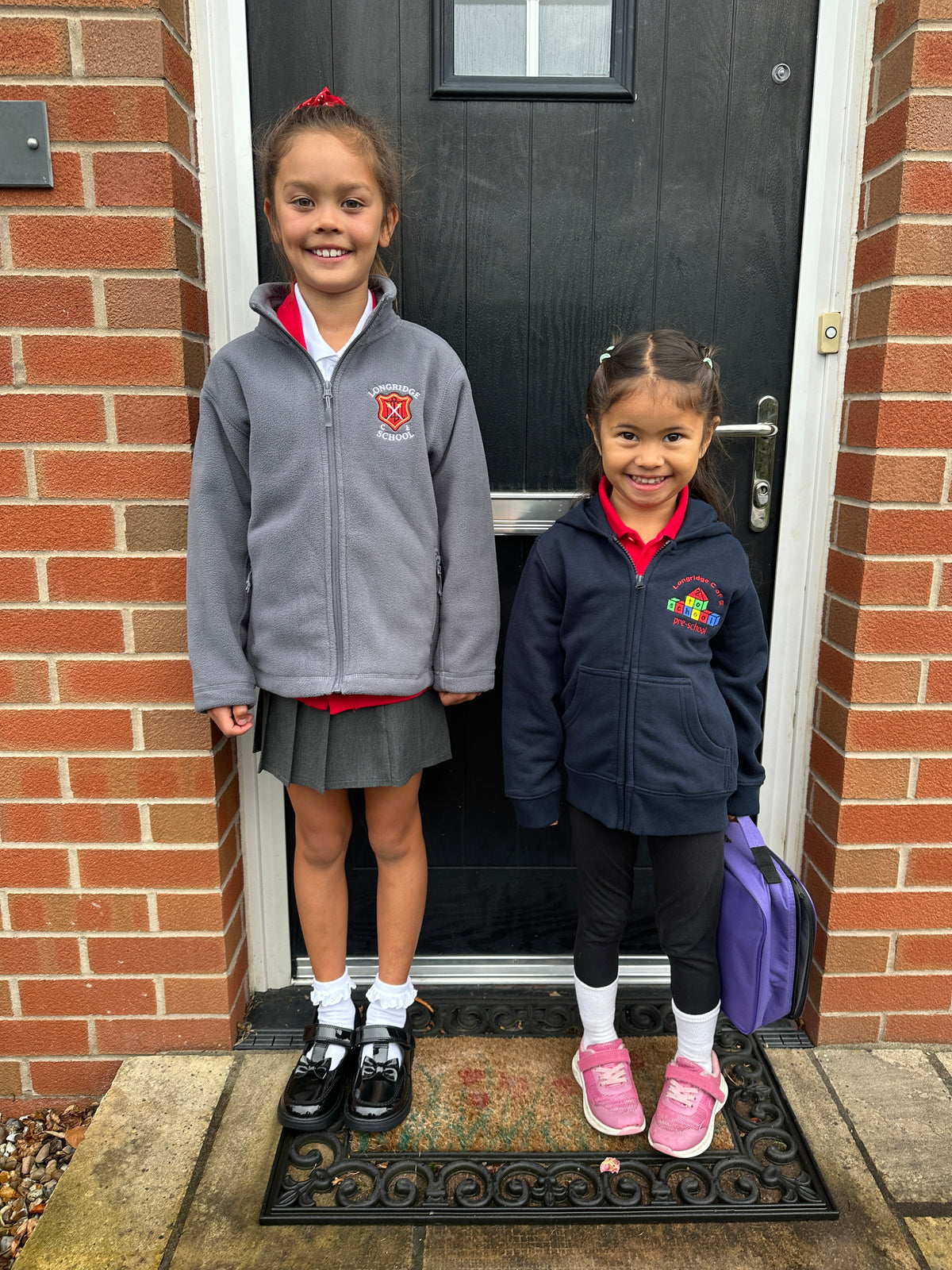 Two young girls standing on a black front door mat in front of a brick house. The girl on the left wears a gray zip-up school jacket with a red and white crest, a gray pleated skirt, white socks, and black shoes.