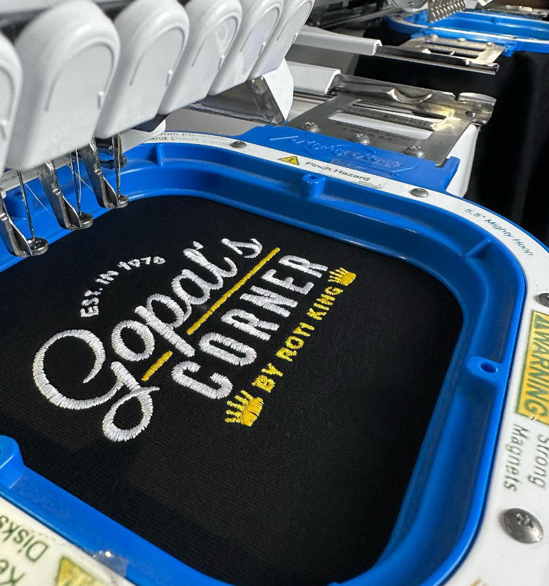 Close-up of an embroidery machine holding a black fabric piece with white and yellow embroidered text reading 'Gopat's Corner' and 'EST. 1975 BY ROTT KING'.