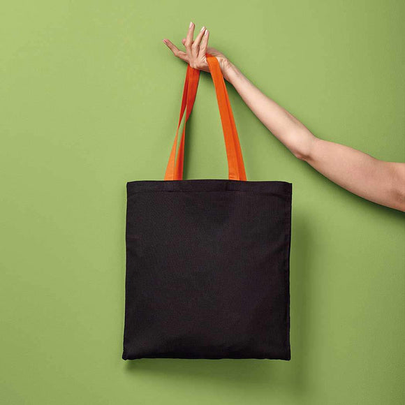 Black tote bag with orange handles held by a hand against a green background