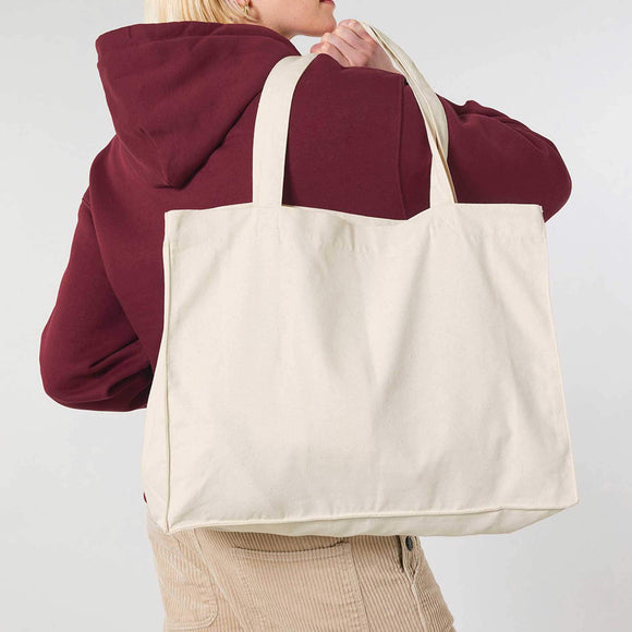 Person wearing a maroon hoodie and beige pants holding a beige tote bag against a white background