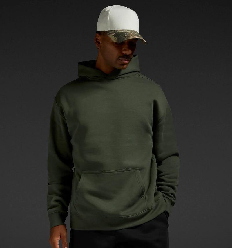 A man wearing an olive green pullover hoodie with a front kangaroo pocket, paired with a white and camouflage baseball cap, set against a dark gray studio background.
