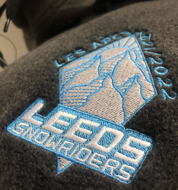 Close-up of an embroidered patch on a dark gray fleece garment. The patch features a light blue and white mountain graphic with the text 'LES APRES SKIING 2022' and 'LEEDS SNOWRIDERS'.
