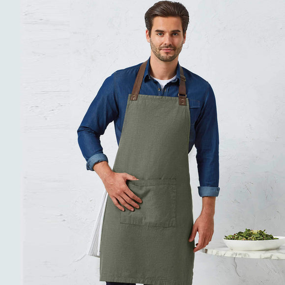 Man wearing a green apron over a blue shirt against a white brick wall.
