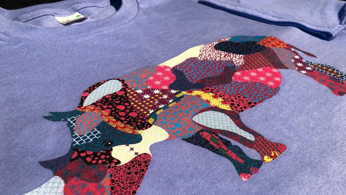 A close-up of a lavender-purple t-shirt featuring a large, colorful patchwork bull graphic on the front. The bull design is composed of various patterned fabric sections in red, pink, black, yellow, and cream.